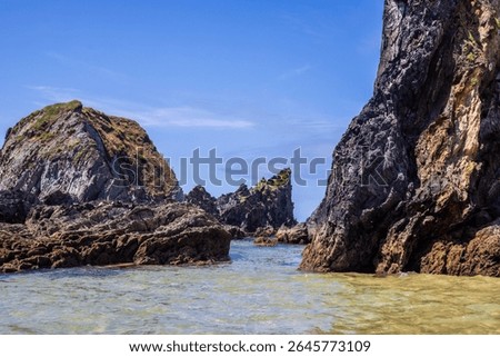 Dramatic rugged rocks frame a sandy beach and clear blue-green ocean under a bright sky. This wild coastal landscape evokes hidden coves and the untouched beauty of nature. Ideal for travel content