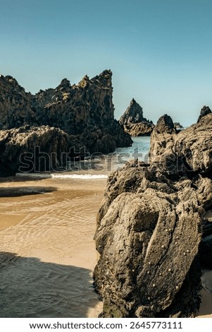 Dramatic rugged rocks frame a sandy beach and clear blue-green ocean under a bright sky. This wild coastal landscape evokes hidden coves and the untouched beauty of nature. Ideal for travel content