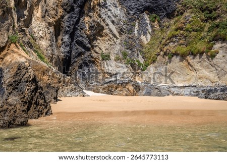 Dramatic rugged rocks frame a sandy beach and clear blue-green ocean under a bright sky. This wild coastal landscape evokes hidden coves and the untouched beauty of nature. Ideal for travel content
