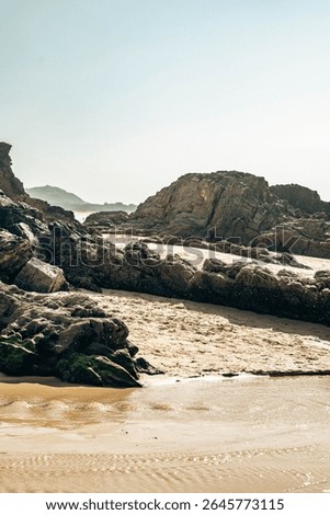 Dramatic rugged rocks frame a sandy beach and clear blue-green ocean under a bright sky. This wild coastal landscape evokes hidden coves and the untouched beauty of nature. Ideal for travel content