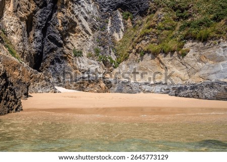 Dramatic rugged rocks frame a sandy beach and clear blue-green ocean under a bright sky. This wild coastal landscape evokes hidden coves and the untouched beauty of nature. Ideal for travel content