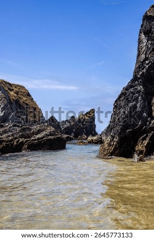 Dramatic rugged rocks frame a sandy beach and clear blue-green ocean under a bright sky. This wild coastal landscape evokes hidden coves and the untouched beauty of nature. Ideal for travel content