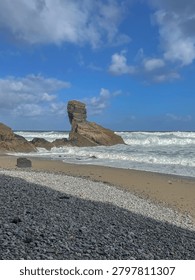 Dramatic sandstone sea stack rising from the pebble and sand beach of Playa de Portizuelo, with crashing waves of the Cantabrian Sea under a blue sky with clouds, Asturias, Spain.