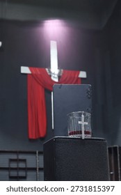 A dramatic scene featuring a white cross draped with red cloth, a bible, and a glass of red liquid with a cross pendant, evoking themes of spirituality and sacrifice