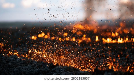 A dramatic scene of a field engulfed in flames, with embers and debris flying through the air against a dusky sky.