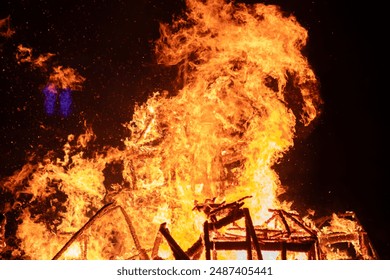 Dramatic scene of a house engulfed in intense flames in a dark night sky. The fire rages, engulfing the structure completely, creating a powerful image of destruction.