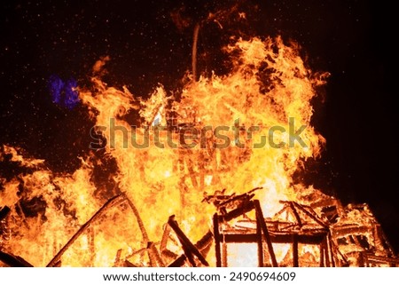Dramatic scene of a house engulfed in intense flames in a dark night sky. The fire rages, engulfing the structure completely, creating a powerful image of destruction.