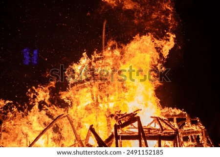 Dramatic scene of a house engulfed in intense flames in a dark night sky. The fire rages, engulfing the structure completely, creating a powerful image of destruction.