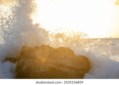 A dramatic scene of a large rock being battered by powerful waves as the sun sets. The sky is ablaze with vibrant colors, reflecting in the crashing water. The scene captures the raw power of nature