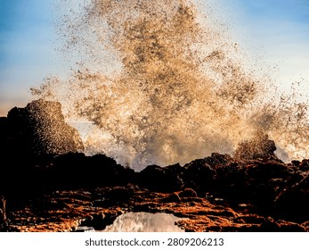A dramatic scene of a massive wave exploding into a spray of water against a rocky shoreline during golden hour