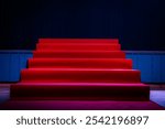 Dramatic scene with a red carpet staircase leading up to a stage, illuminated by blue spotlights. The stage is set for an award show, performance, or presentation.