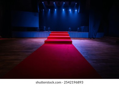 Dramatic scene with a red carpet staircase leading up to a stage, illuminated by blue spotlights. The stage is set for an award show, performance, or presentation.