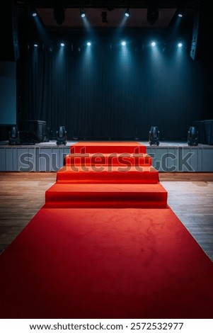 Dramatic scene with a red carpet staircase leading up to a stage, illuminated by blue spotlights. The stage is set for an award show, performance, or presentation.