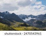 Dramatic scenery in alpine valley with creek among green hills and rocks with view to rocky pointy peak, large snow-capped peaked top, mountain range and big glacier tongue under clouds in blue sky.