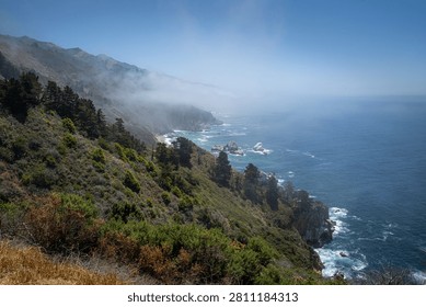 Dramatic scenic view of the rugged Big Sur coastline along Highway 1 with a rolling marine layer fog bank hitting the green ocean cliffs, California.