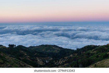 Dramatic sea of clouds over Madeira's mountainous landscape. Aerial view of lush green hills meeting dense fog. Atlantic Ocean horizon in the distance. Volcanic island scenery at sunrise.