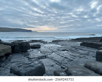 A dramatic sea scape at dusk, featuring waves crashing against rugged black volcanic rock along the shoreline.