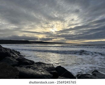 A dramatic sea scape at dusk, featuring waves crashing against rugged black volcanic rock along the shoreline.