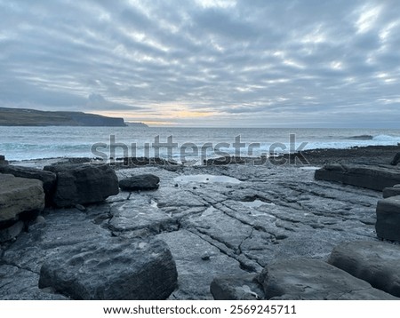 A dramatic sea scape at dusk, featuring waves crashing against rugged black volcanic rock along the shoreline.