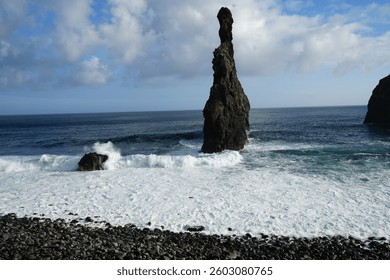 A dramatic sea stack rises from the ocean, surrounded by crashing waves and deep blue waters under a sky with fluffy clouds, creating a striking and powerful coastal landscape.