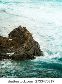 Dramatic seascape captures a rugged basalt rock formation battling relentless turquoise waves. Foamy sea spray highlights erosion patterns on the stone, illustrating nature's raw power.