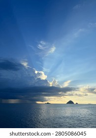 Dramatic seascape with crepuscular sun rays shining through clouds over the ocean, rain falling in the distance with small islands on the horizon during golden hour

