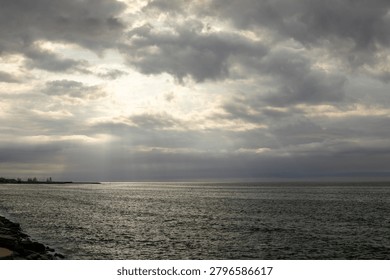 Dramatic seascape featuring dark storm clouds over a calm ocean with rays of sunlight breaking through the cloud cover and illuminating the water surface. Atmospheric coastal landscape with moody weather, natural light, open horizon, and tranquil marine scenery.