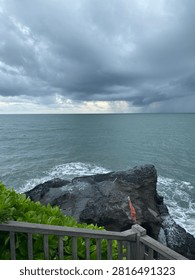 A dramatic seascape featuring dark storm clouds gathering over the ocean with visible rainfall in the distance. Waves crash against a rugged rocky shoreline while lush green coastal plants frame the foreground, creating a powerful natural landscape ideal for travel, weather, climate, and nature concepts.