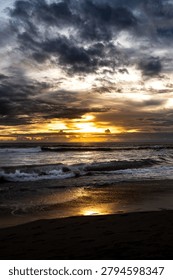 A dramatic seascape featuring powerful ocean waves beneath dark storm clouds at sunset. Golden sunlight breaks through the cloud-covered horizon, creating beautiful reflections on the wet sandy beach. The contrast between the moody sky and warm sunset tones captures the raw beauty and atmosphere of a coastal landscape