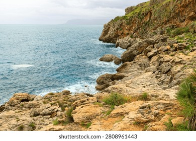 A dramatic seascape featuring weathered rock formations meeting the vibrant blue ocean with white foam