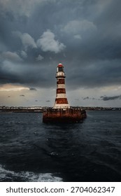 Dramatic seascape with an old lighthouse standing alone in the water under dark storm clouds. Moody coastal scene with rough waves, historic tower and distant shoreline at sunset light.