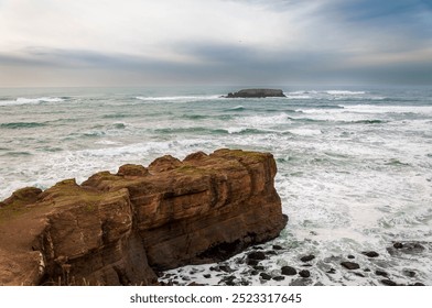 Dramatic seascape with rocky cliffs and a distant island under a cloudy sky.
