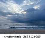 Dramatic seascape with storm clouds gathering over the ocean horizon and gentle waves washing ashore on a sandy beach.