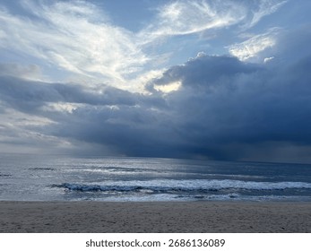 Dramatic seascape with storm clouds gathering over the ocean horizon and gentle waves washing ashore on a sandy beach.