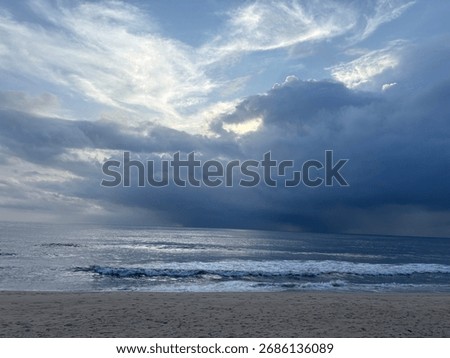 Dramatic seascape with storm clouds gathering over the ocean horizon and gentle waves washing ashore on a sandy beach.