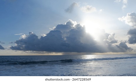 A dramatic seascape as the sun breaks through a large storm cloud, creating a silver lining and sunbeams over the ocean. A distant rain shower is visible above the calm sea.
