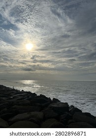 Dramatic seascape with sunlight breaking through textured clouds over a calm ocean, reflecting on the water and rocky shoreline, creating a peaceful coastal nature scene.