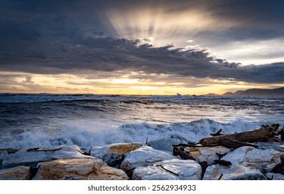 A dramatic seascape at sunset, featuring turbulent waves crashing against rocky shorelines. Dark clouds loom overhead, with rays of sunlight breaking through, creating a stunning contrast in the sky. - Powered by Shutterstock