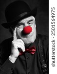 Dramatic selective color portrait of a sad clown against a dark background. An elderly man with red clown nose and bowler hat on head. Dressed in black shirt with suspenders. Hand near face.