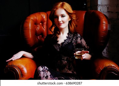 Dramatic Sensual Redhead Woman With Blue Eyes In Black Lace Peignoir Drinking Whiskey Sitting Comfortably In Vintage Leather Armchair