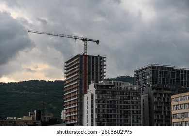 A dramatic shot capturing a tall construction crane towering over a partially completed high-rise apartment building. The scene is set against a backdrop of dense, dark storm clouds and a green, forested hillside in the city.