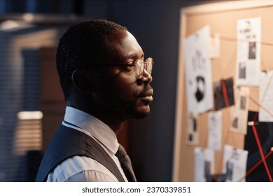 Dramatic side view portrait of Black male detective looking at evidence board in office while investigating case