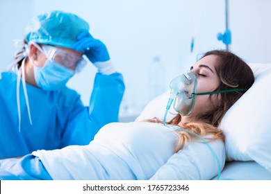 Dramatic Side View Of A Young Female Patient Breathing Assisted By A Respiratory Tube And An Exhausted Dedicated Doctor In The Intensive Care Unit Of A Hospital