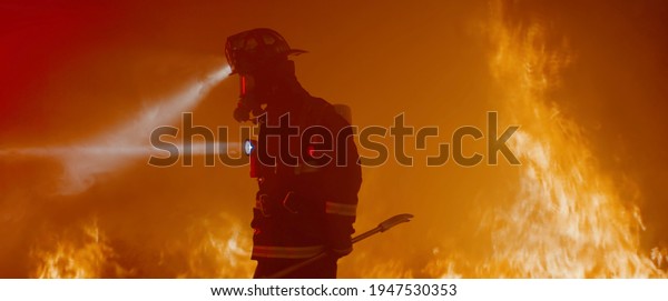 Dramatic silhouette of American firefighter in\
full gear exploring the huge fire\
zone