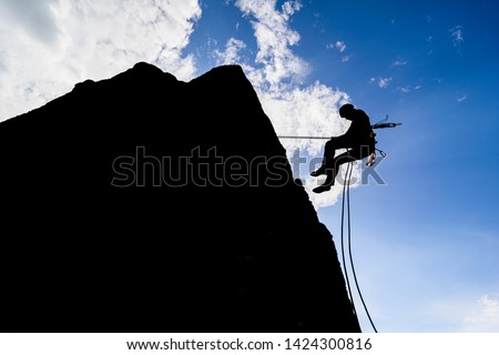A dramatic silhouette of a climber rappeling down a rock wall. Rock climber with a rope abseil down. Mountaineer jumping down on climbing rope. Extreme adventure sport of rock climbing.