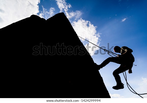 A dramatic silhouette of a climber rappeling\
down a rock wall. Rock climber with a rope abseil down. Mountaineer\
jumping down on climbing rope. Extreme adventure sport of rock\
climbing.