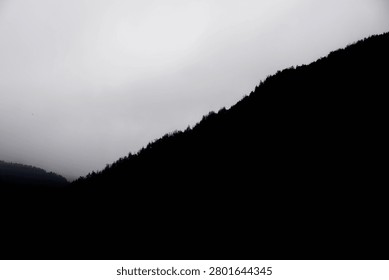 A dramatic silhouette of a forested mountain slope against a bright, overcast, and hazy sky.