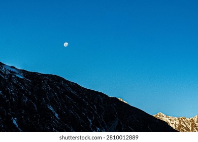 Dramatic silhouette of a rugged mountain range against a vibrant deep blue sky with a bright moon. The dark rocky peaks create a powerful and serene composition, capturing the majestic beauty of New Zealand’s Southern Alps during twilight.