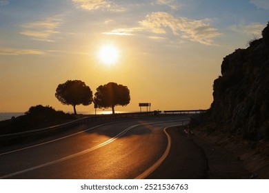 A dramatic silhouette of trees against the sunset sky on a coastal road overlooking the Mediterranean in Finike, highlighting peaceful evening hues.