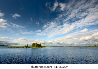 Dramatic sky with cirrus and cumulus clouds above the blue lake and island with green birch trees before the rain. Idyllic summer scene. Panoramic view. Vacations, ecotourism, nature of Scandinavia